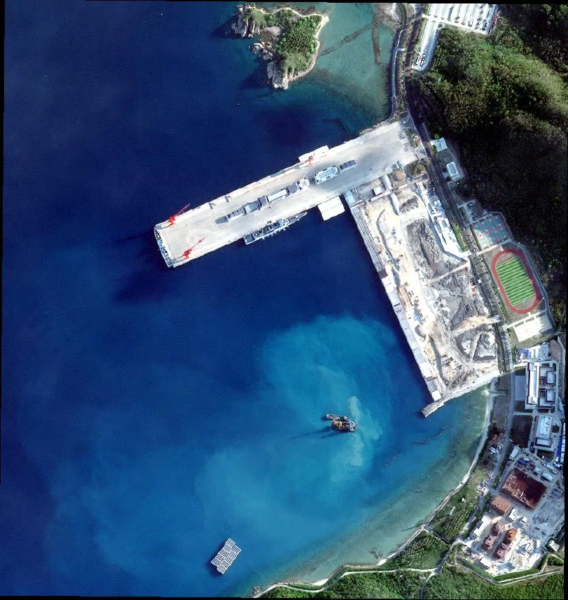 Satellite image of a pier at the Yulin naval port on Hainan Island. A long concrete pier with two red cranes extends into deep blue water. Gray vessels are moored along its south side. The adjoining quay is a construction site with excavated ground. A sports field and buildings sit on shore, and a small vessel and a floating structure are in the bay